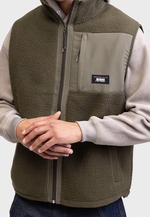 Mężczyzna w oliwkowej kamizelce polarowej z zamkiem i kieszenią na piersi, noszący ją na jasnobeżowej bluzie, z rękami złożonymi na przodzie.