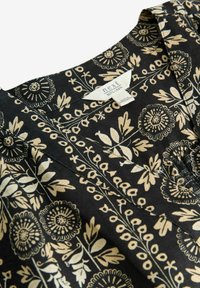 Tissu en lin noir avec un motif floral beige, présentant un design en v et des plis à l'avant. L'étiquette indique "next, 100% lin."