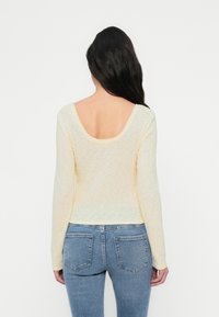 Μακρυμάνικη, ριμπ μπλούζα σε ανοιχτό κίτρινο χρώμα με φαρδιά στρογγυλή πλάτη. Συνδυάζεται με μπλε τζιν παντελόνι, αποκαλύπτοντας λεπτομέρειες στην πλάτη και εφαρμοστή σιλουέτα.