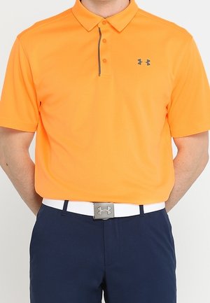 Man draagt een feloranje poloshirt met korte mouwen, ingestopt in marineblauwe broek met een witte Under Armour-riem.
