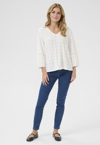 Haut en coton blanc avec un motif en œillets, décolleté en V et manches trois-quarts, assorti à un jean ajusté bleu et des ballerines noires.