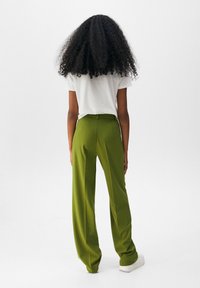 Grüne weit geschnittene Hose mit glatter Textur und maßgeschneiderter Passform, kombiniert mit einem figurbetonten weißen T-Shirt. Das Modell wird von hinten betrachtet.