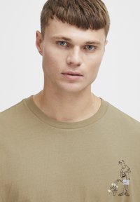 Olivengrøn bomulds t-shirt med korte ærmer og rund hals. Har et cartoon grafisk billede af en person, der vander en plante på venstre bryst.
