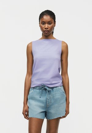 Kvinde med flettet hår, iført en ærmeløs lavendelfarvet top og lyseblå denimshorts med bindebånd i taljen, stående mod en ensfarvet baggrund.