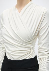 Femme portant un top blanc à manches longues froncé et cache-cœur, associé à un pantalon noir taille haute, montrée du cou à la taille.