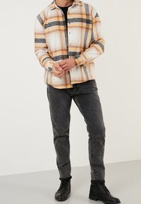 Camisa de franela a cuadros en crema, naranja y azul marino, con botones al frente y mangas largas. Combinada con vaqueros grises y botas negras con cordones.