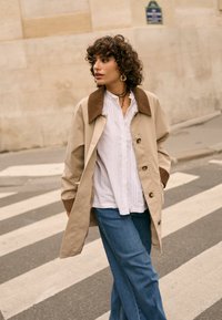 Osoba o kręconych włosach, ubrana w beżowy płaszcz, białą bluzkę i niebieskie dżinsy, przechodzi przez przejście dla pieszych w mieście, patrząc na bok.