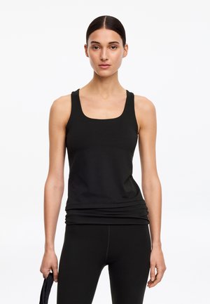 Jeune femme aux cheveux foncés attachés, portant un débardeur noir et des leggings noirs, se tenant devant un fond blanc uni.