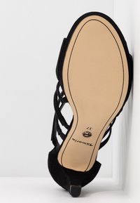 Sandale à talons hauts en daim noir avec un design à lanières, présentant une semelle beige avec un léger talon et un accent circulaire sur le dessous. Taille 37.