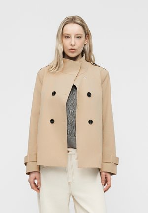 Femme portant une veste beige croisée à boutons noirs sur un pull en maille torsadée gris et un pantalon blanc cassé, debout devant un fond uni.