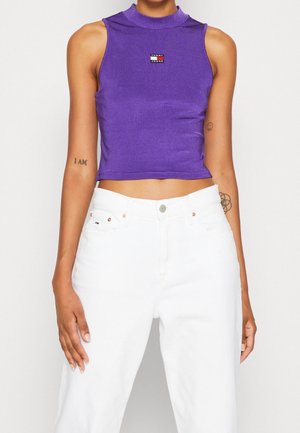 Osoba w bezrękawniku w kolorze fioletowym z logo "Tommy Jeans" oraz wysokim stanem białych spodni, ukazująca tatuaże na ramionach i przyjmująca swobodną pozę.