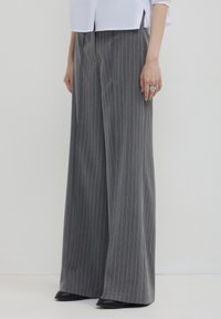 Frau in grauen, gestreiften, weiten Hosen, schwarzen Schuhen und einer weißen Bluse, die vor einem einfarbigen, hellen Hintergrund steht.