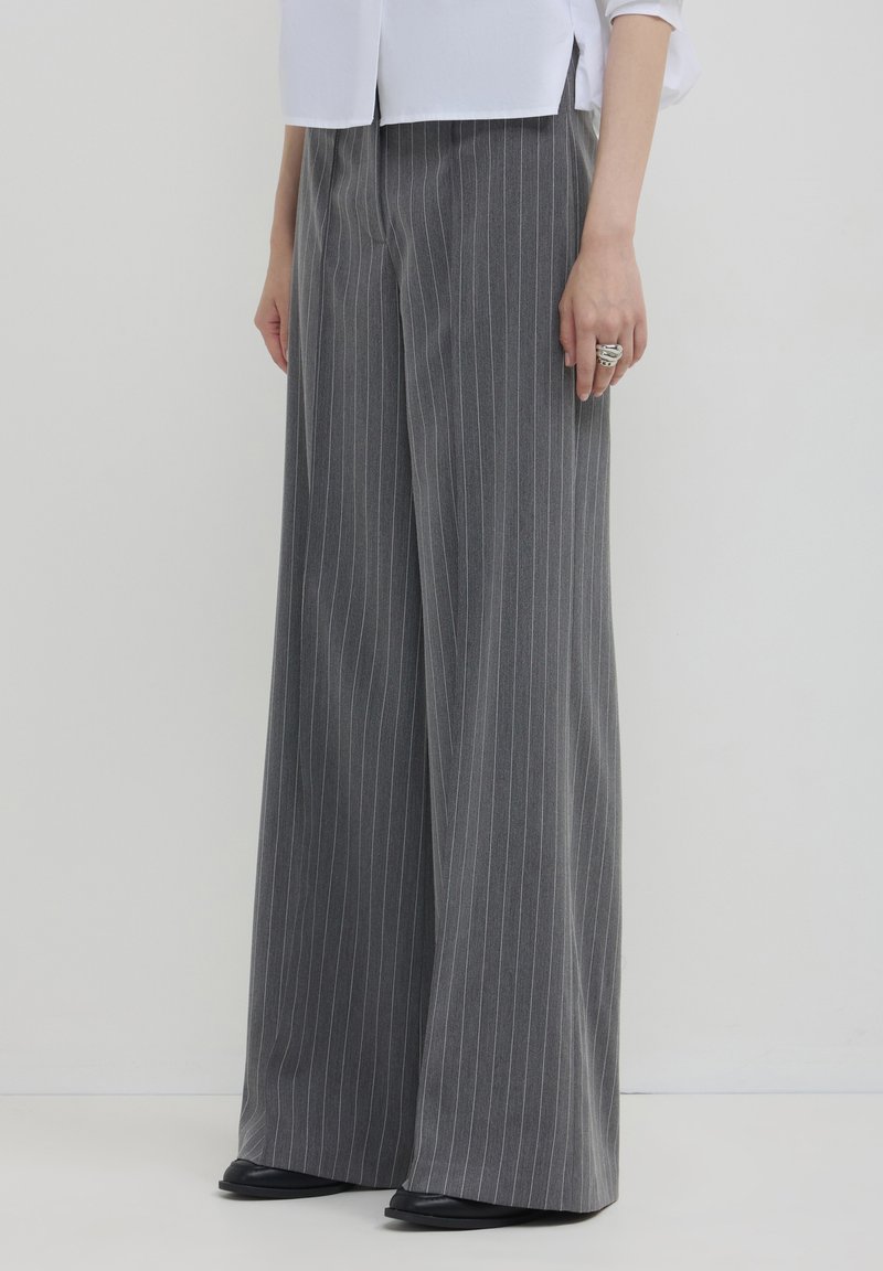 Frau in grauen, gestreiften, weiten Hosen, schwarzen Schuhen und einer weißen Bluse, die vor einem einfarbigen, hellen Hintergrund steht.