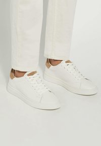White leather sneakers with a smooth texture and light brown suede accents at the heel. Features a rounded toe and flat rubber sole.