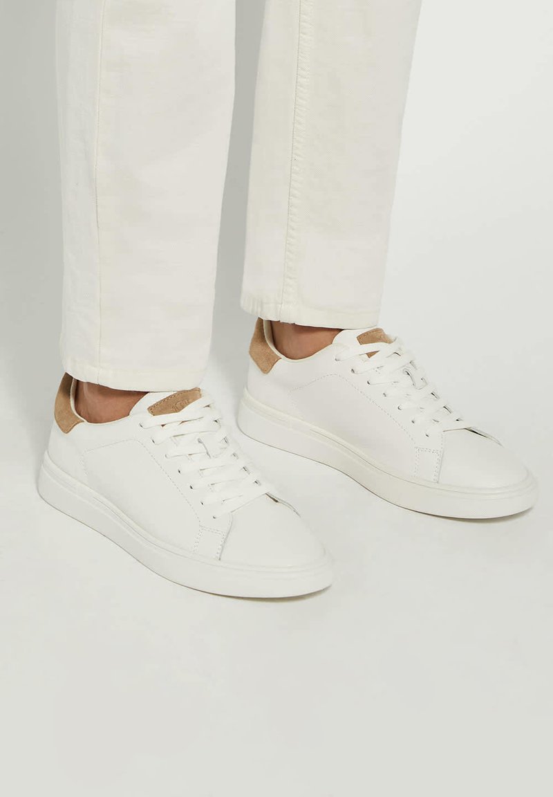 White leather sneakers with a smooth texture and light brown suede accents at the heel. Features a rounded toe and flat rubber sole.
