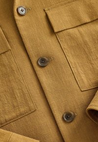 Camisa de lino marrón con dos cierres de botón, bolsillo de parche y una superficie texturada. Presenta botones suaves y redondeados. Tejido suave y ligero.