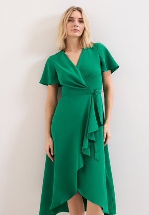 Femme aux cheveux blonds mi-longs portant une robe portefeuille verte à manches courtes avec un détail volanté sur la jupe.