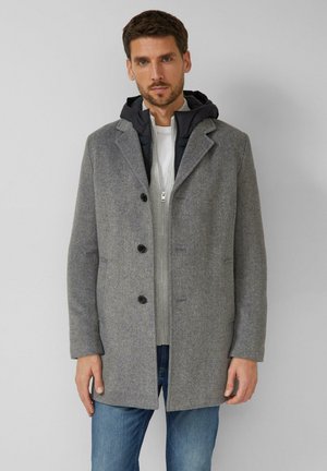 Mann trägt einen grauen, geknöpften Wollmantel über einer schwarzen Kapuzenjacke und einem hellgrauen Reißverschluss-Pullover, blaue Jeans, steht vor einem einfarbigen Hintergrund.