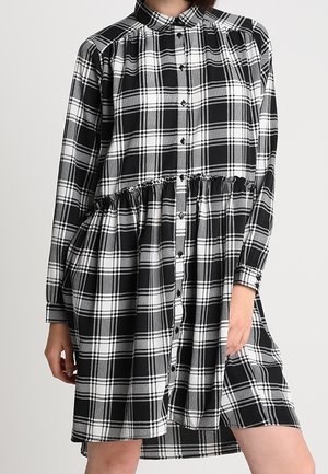 Femme portant une robe chemise à manches longues à carreaux noirs et blancs avec un détail volanté à la taille, debout devant un fond uni.