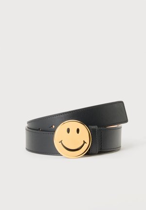 Ceinture en cuir noir roulée avec une grande boucle ronde dorée représentant une icône de visage souriant sur un fond clair uni.