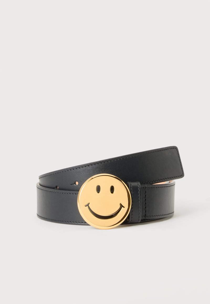 Ceinture en cuir noir roulée avec une grande boucle ronde dorée représentant une icône de visage souriant sur un fond clair uni.