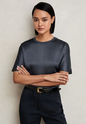 Mujer con cabello liso y negro, vestida con una camiseta corta de satén oscuro y pantalones negros con un cinturón negro, de pie con los brazos cruzados.