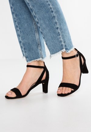 Stopy w jasnoniebieskich, postrzępionych dżinsach, ubrane w czarne zamszowe sandały na blokowym obcasie z kostką, na białej powierzchni.