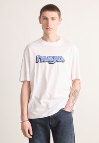 White cotton t-shirt featuring a blue patterned "hugo." logo, short sleeves, relaxed fit, paired with dark blue jeans and silver bracelet.