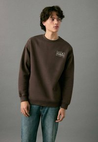 Bruine sweater met een ronde hals en geribbelde manchetten. Voorzien van een wit logopatch op de borst. Gecombineerd met lichtblauwe spijkerbroek.