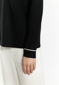 Zwarte trui met een zachte textuur; heeft een witte accent op de manchetten. Gepaard met lichtgekleurde broeken, die het contrast van de outfit benadrukken.