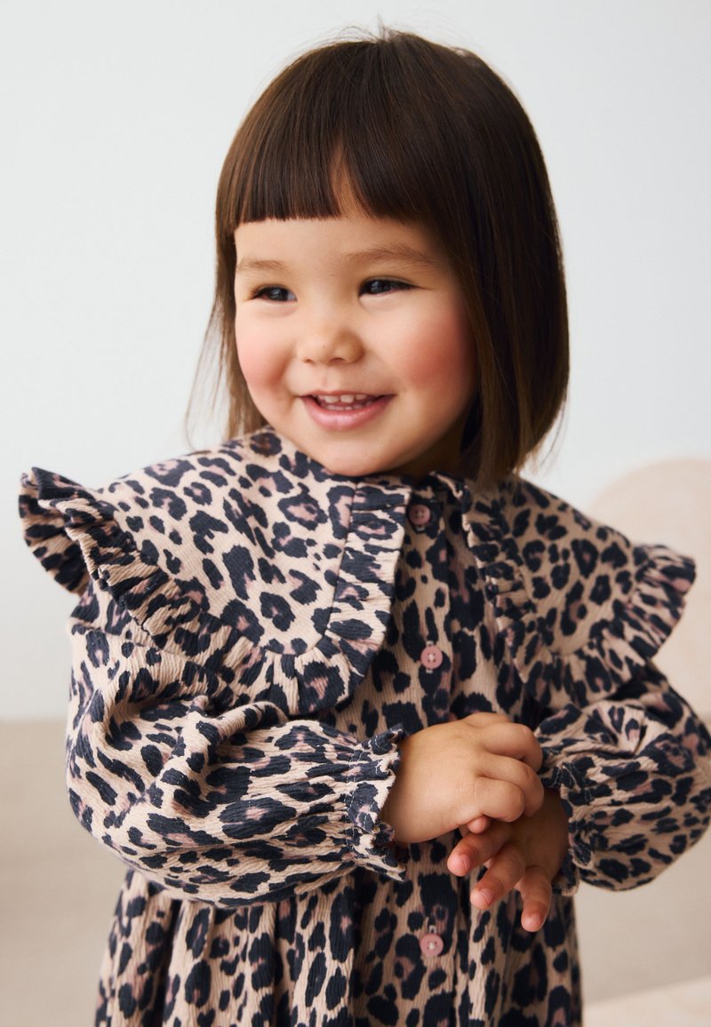 Smilende ungt barn med glat mørkt hår iført en langærmet kjole med leopardprint og flæser på skuldrene, stående med sammenfoldede hænder.