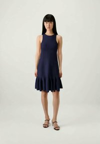 Mulher jovem com cabelo preto longo, vestindo um vestido sem mangas azul escuro com bainha em babados e sandálias de salto alto pretas com tiras, de pé e em pé direito.