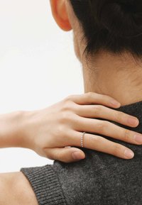 Anillo de plata con una fila de pequeñas piedras preciosas, llevado en una mano descansando sobre un suéter gris oscuro, mostrando piel suave y uñas cortas.
