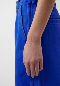 Blå denimbyxor har en sidoficka med grön söm som accent, visad från sidan, vilket framhäver tygets textur och passform.