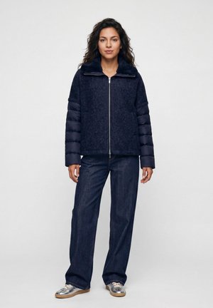 Frau steht und trägt eine marineblaue Jacke mit Reißverschluss und gepolsterten Ärmeln, dunkle weite Jeans und silberne Sneakers vor einfarbigem Hintergrund.