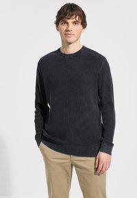 Pull tricoté sombre et texturé avec un col rond, manches longues et poignets côtelés, associé à un pantalon beige clair.