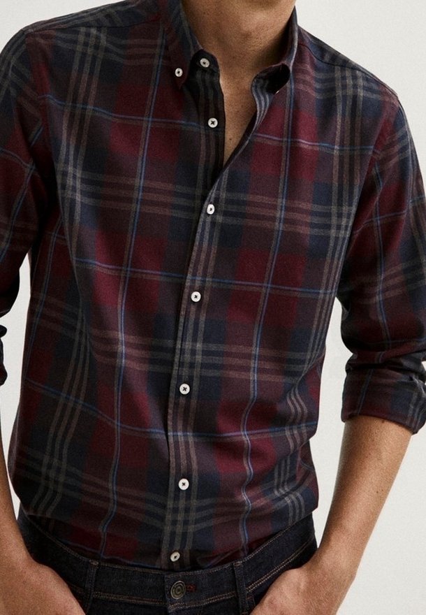 Homme portant une chemise à manches longues à carreaux boutonnée, aux tons rouge foncé, noir, gris et bleu, avec les manches retroussées.