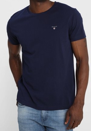 Man wearing navy blue GANT t-shirt and light blue jeans, standing with one hand in pocket against white background.