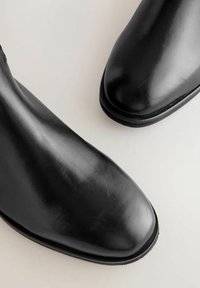 Pari kengät kiillotettua mustaa nahkaa, joissa on pyöreät varpaat, vaaleanharmaalla pinnalla, kuvattu ylhäältä päin.