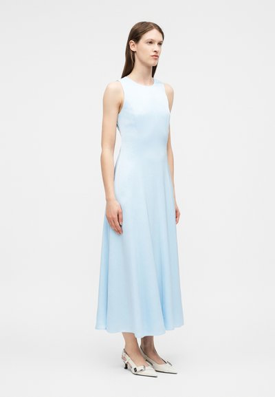 Femme debout vêtue d'une robe midi sans manches bleu clair et de chaussures blanches à talons bas et bouts pointus avec boucles.