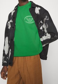 Giacca tie-dye bianca e nera sopra una maglietta verde con un logo in rilievo, abbinata a pantaloni marrone strutturati.