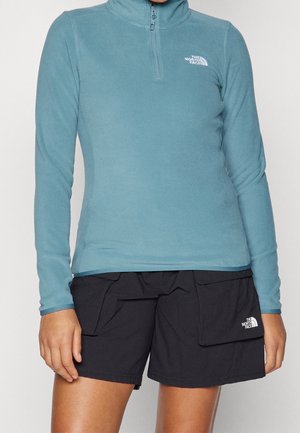 Kvinde iført en lyseblå langærmet top med lynlås ved halsen og sorte cargoshorts, begge med The North Face-logo.