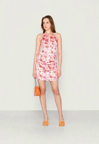 Jovem mulher com um vestido mini halter em flores cor-de-rosa e vermelhas, segurando uma mala laranja, vestindo sandálias de salto laranja, em pé contra um fundo simples.