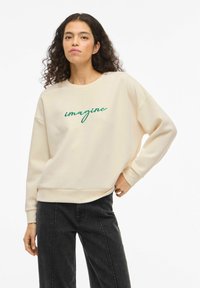 Kremfarget genser laget av mykt stoff, med grønn brodert tekst "imagine." Løs passform med ribbestrikkede mansjetter og rund hals.