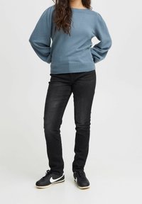 Jasnoniebieski sweter z dzianiny z szerokim dekoltem i bufkami na rękawach, zestawiony z czarnymi, dopasowanymi jeansami oraz czarnymi sneakersami z białymi akcentami.