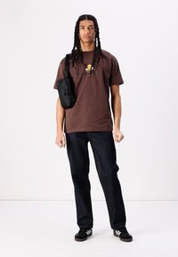 Brun bomulls t-skjorte med grafisk design, kombinert med mørk denim jeans. En svart crossover-veske og sneakers fullfører antrekket.