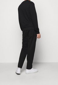 Uomo che indossa una felpa nera e pantaloni neri con sneakers bianche, in piedi contro uno sfondo grigio chiaro uniforme, visto da dietro.