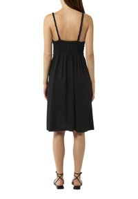 Schwarzes Kleid aus weichem Stoff, mit dünnen Trägern, einem gerafften Oberteil und einem knielangen Rock. Getragen mit schwarzen Riemchensandalen.