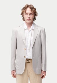Veste grise claire avec deux poches avant, revers crantés et deux boutons. Portée avec une chemise blanche et un pantalon beige.