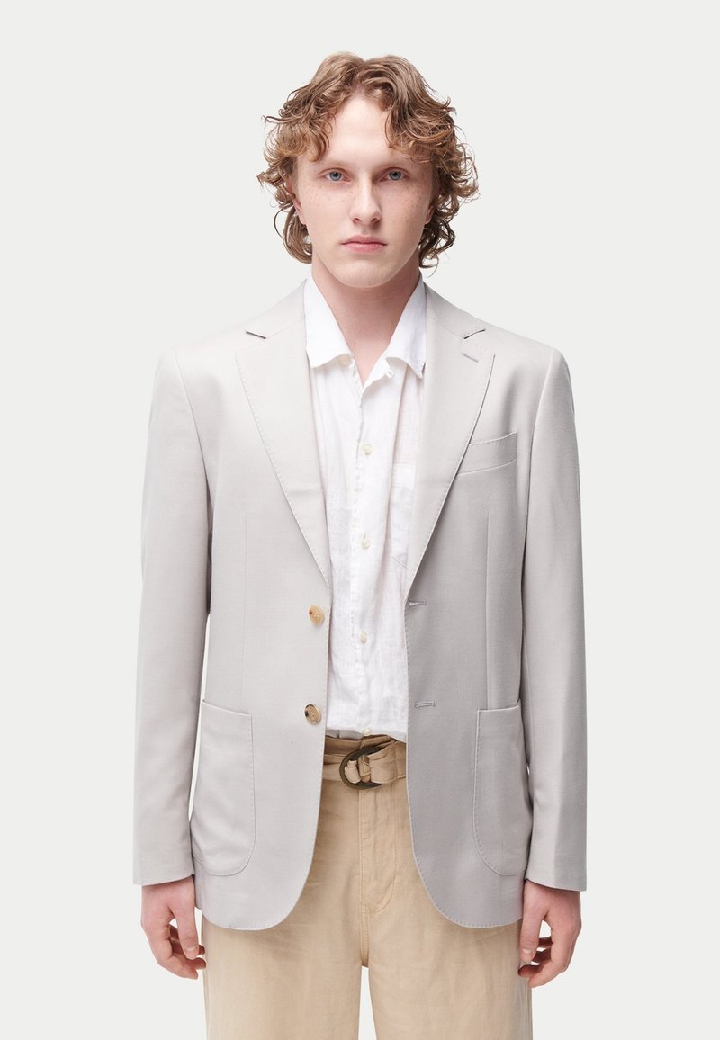 Veste grise claire avec deux poches avant, revers crantés et deux boutons. Portée avec une chemise blanche et un pantalon beige.